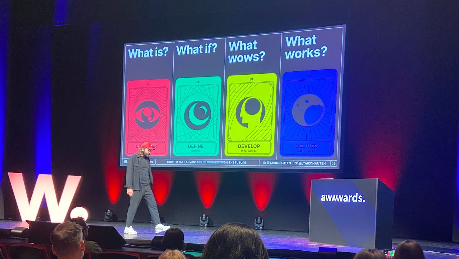 The image shows a speaker on stage at the Awwwards event. In front of him is a large screen with three colorful panels, posing the questions "What is?", "What if?" and "What wows?", each accompanied by a corresponding word: DISCOVER, DEFINE, and DEVELOP, 