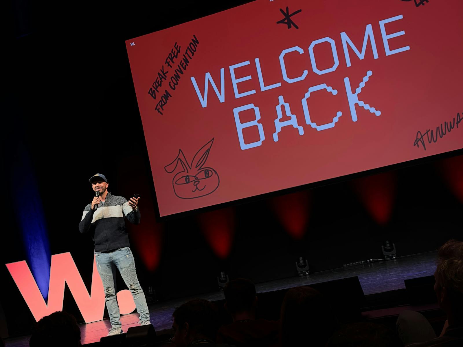 De afbeelding toont een spreker op een podium tijdens het Awwwards evenement. Achter de spreker staat een groot scherm met de tekst 'WELCOME BACK' prominent weergegeven in witte letters op een rode achtergrond. De sfeer is informeel en de spreker draagt c