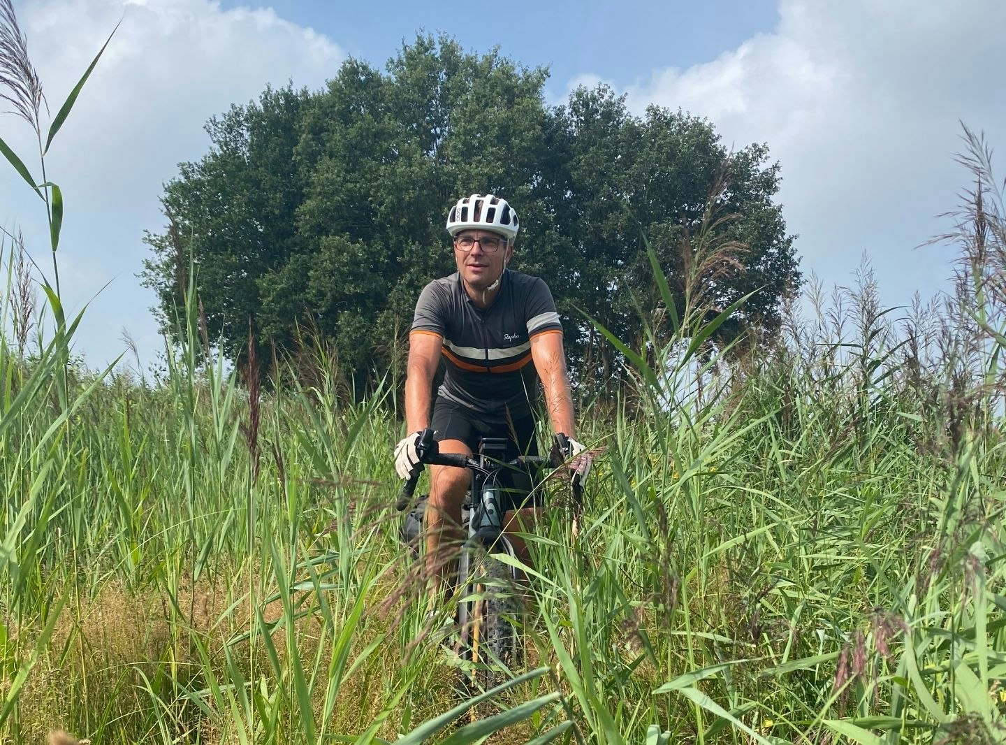 Persoon fietsend door hoog gras, met een grote boom op de achtergrond.