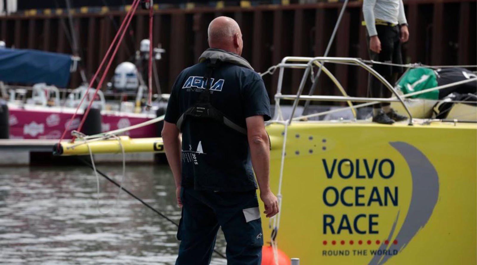 Man die vanaf het water naast een Volvo Ocean Race bord staat met rug naar de toeschouwer